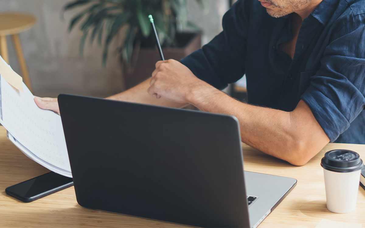 Mann sitzt vor einem Laptop mit Zettel in der Hand am Tisch