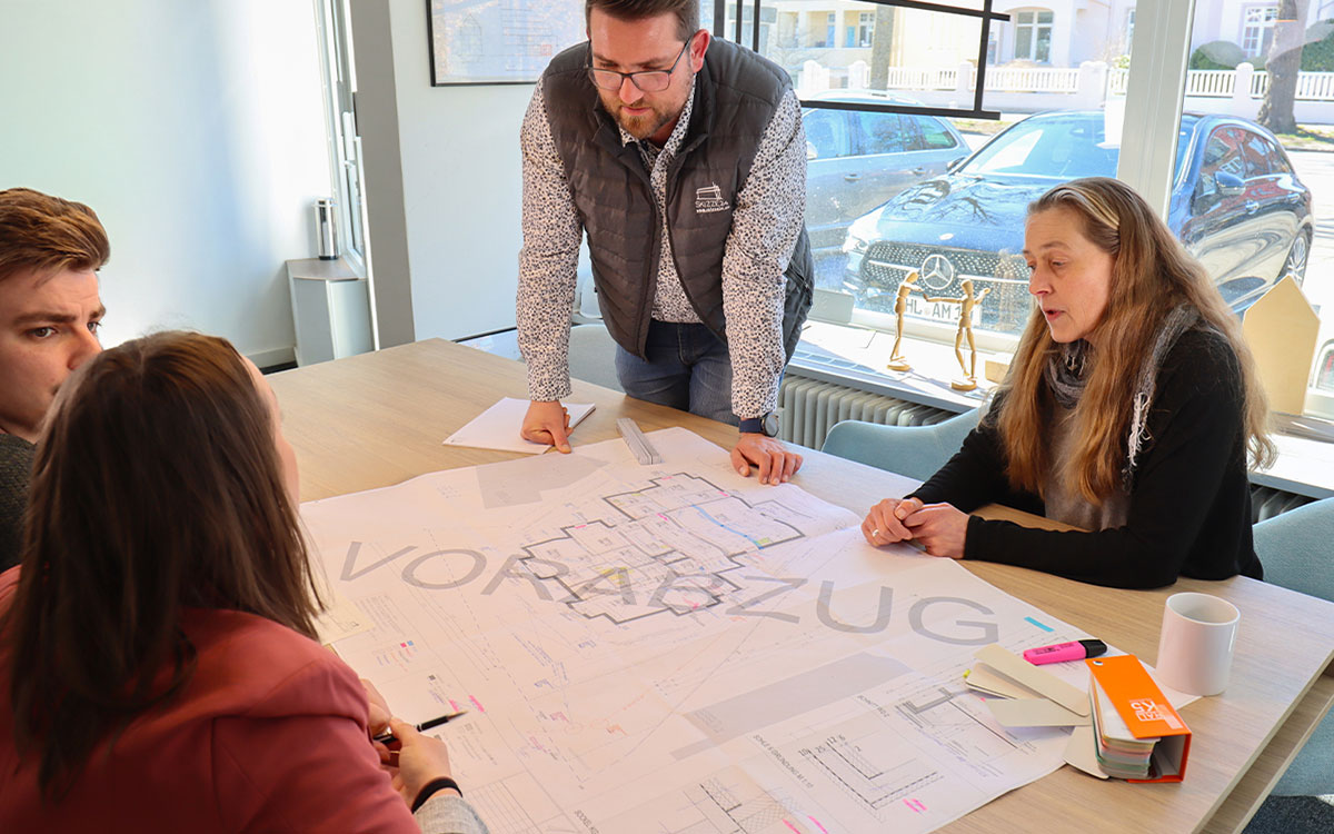Vier Personen sitzen um einen Konferenztisch und besprechen Baupläne in einem Büro. Ein Mann steht und erklärt Details auf einem Architekturplan.
